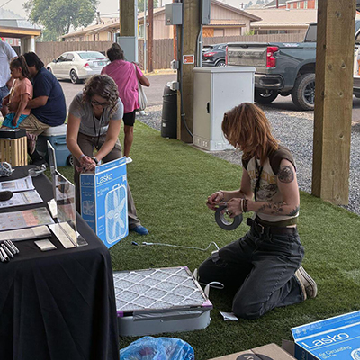 Ariel Cowan (background, middle) and Jenna Deibel (far right) assemble box fan air filters at the Thursday outdoor market in Warm Springs.