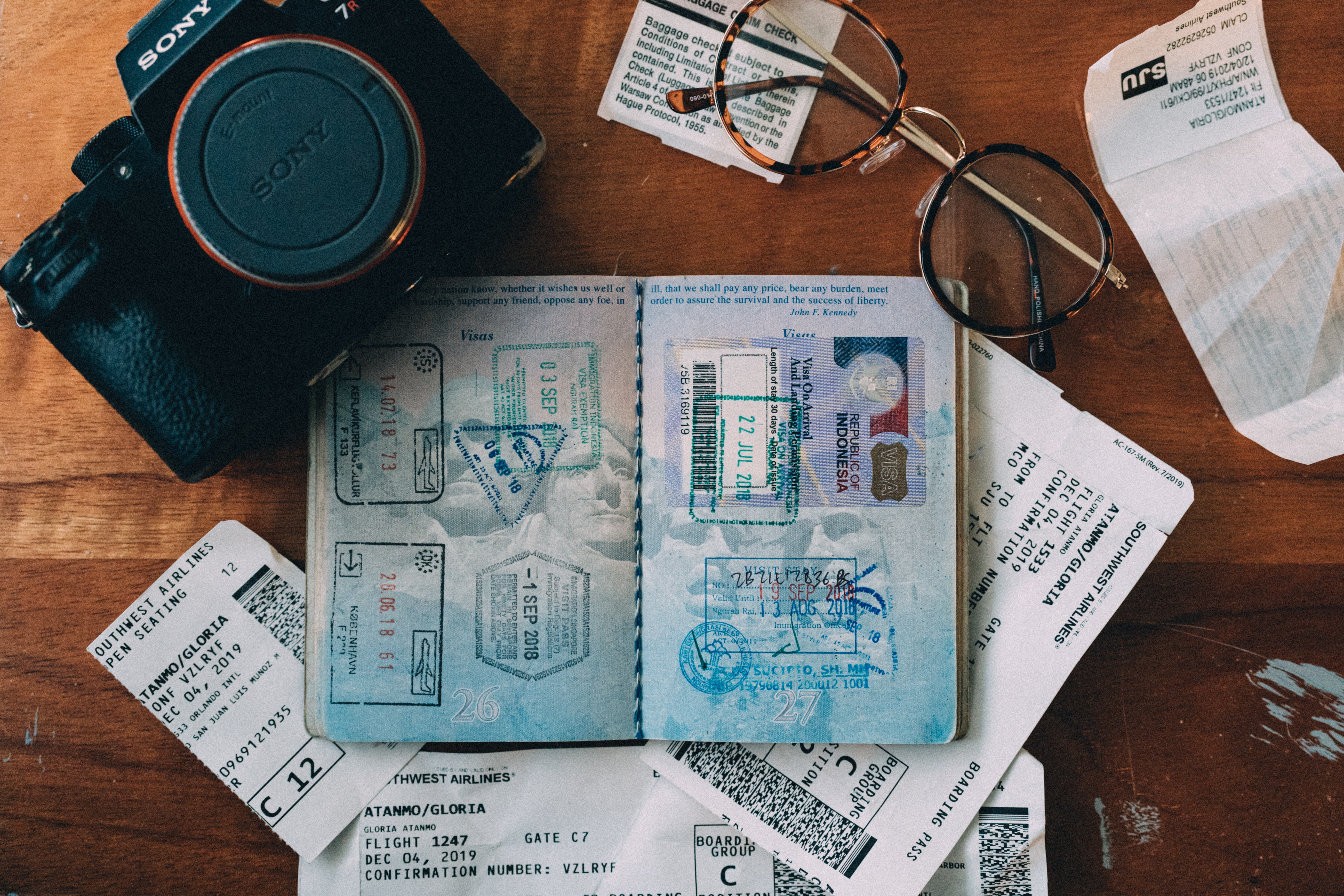 A passport with many stamps rests on a table. There is a camera, a pair of glasses and some plane tickets on the table.