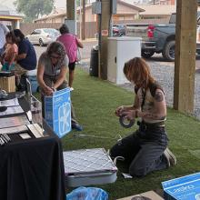 Ariel Cowan (background, middle) and Jenna Deibel (far right) assemble box fan air filters at the Thursday outdoor market in Warm Springs.